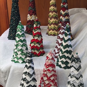May include: Nine handmade fabric Christmas trees in various colors and patterns, including red, green, white, and gold. The trees are arranged on a white tablecloth.