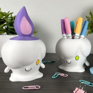 May include: Two white candle-shaped desk organizers with smiling faces and yellow or green eyes. One organizer has a purple flame lid, while the other is open and holds five colorful markers. A separate blue flame lid sits nearby with scattered paper clips.