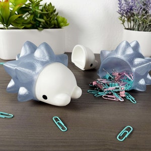 May include: Two blue and white spiky Pokemon-themed desk organizers. One organizer is filled with colorful paper clips. The organizers are on a wooden surface.