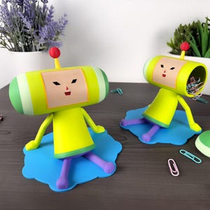 May include: Two whimsical, yellow and green figures with red antenna-like tops and light blue bases. The figures have a cartoonish design with a face on the front. One figure is upright, while the other is tilted, revealing the inside. Paperclips are scattered around.