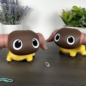 May include: Two cartoon-style figurines of a ram-like creature with brown heads, large white-rimmed eyes, and pink horns. The bodies are yellow. The figurines are on a dark wooden surface, with paperclips and potted plants in the background.