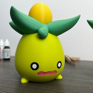 May include: A 3D printed yellow and green figurine with a round body, two small legs, and a large green leaf on top. The figurine has a cute, cartoon-like face with black eyes and a pink mouth.