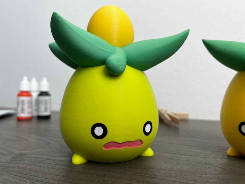 May include: A 3D printed yellow and green figurine with a round body, two small legs, and a large green leaf on top. The figurine has a cute, cartoon-like face with black eyes and a pink mouth.