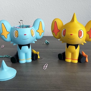 May include: Two Shinx-themed desk organizers, one light blue and one yellow, with open tops and paperclips inside. The organizers have large ears and star-shaped accents. A matching lid sits next to the blue organizer. The organizers are on a gray surface.