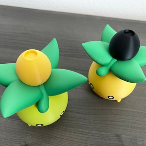 May include: Two yellow and green 3D printed figurines with black tops. The figurines are shaped like a plant with a yellow bulb on top.