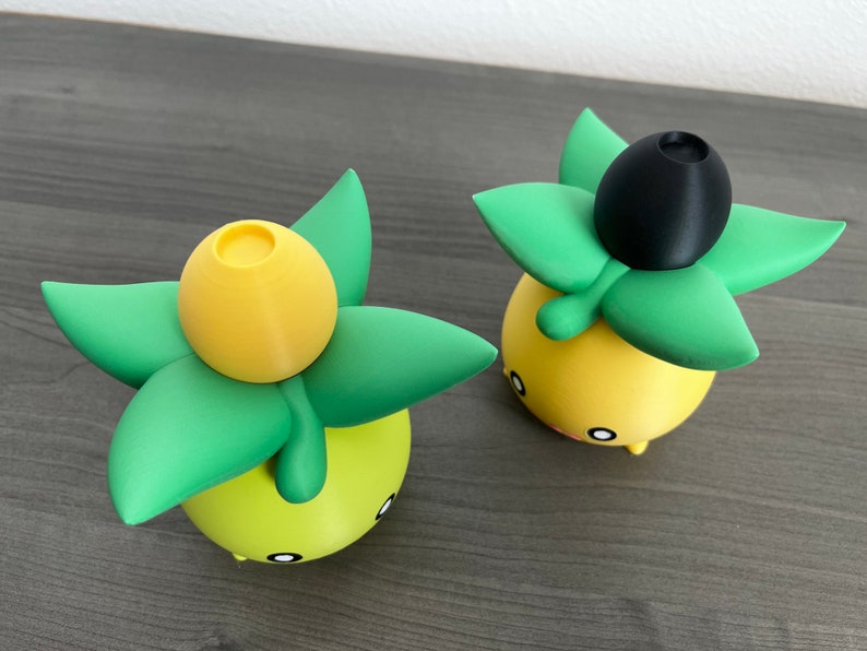 May include: Two yellow and green 3D printed figurines with black tops. The figurines are shaped like a plant with a yellow bulb on top.