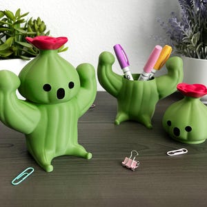 May include: Three green cactus-shaped desk accessories with smooth matte finishes. One large cactus figure with a red flower, one cactus-shaped pen holder containing three markers, and one small cactus figure with a red flower.