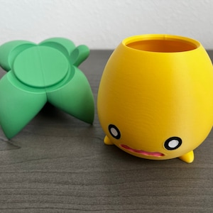 May include: A yellow ceramic planter with a cute face and two small legs. The planter is sitting on a dark wood surface next to a green ceramic leaf-shaped object.