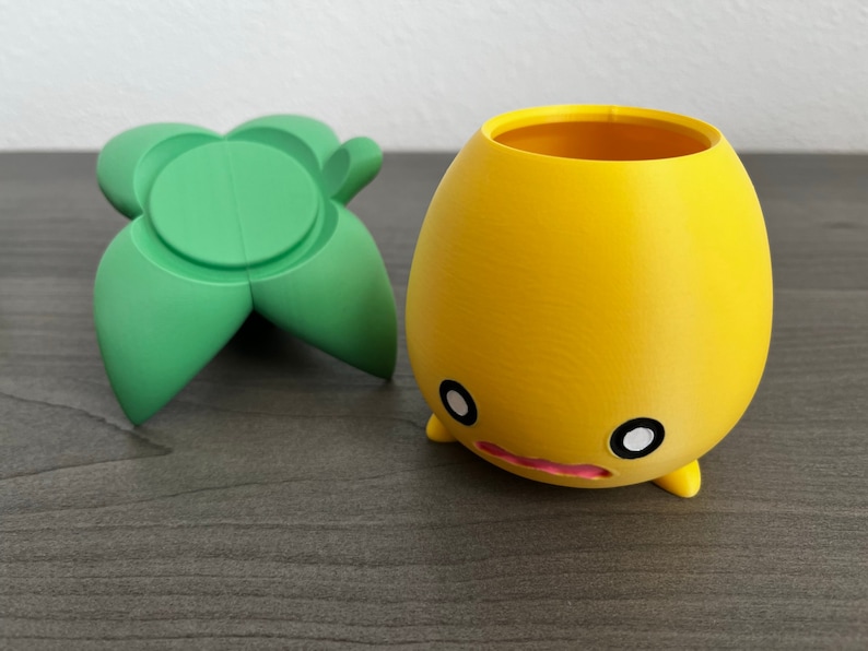 May include: A yellow ceramic planter with a cute face and two small legs. The planter is sitting on a dark wood surface next to a green ceramic leaf-shaped object.