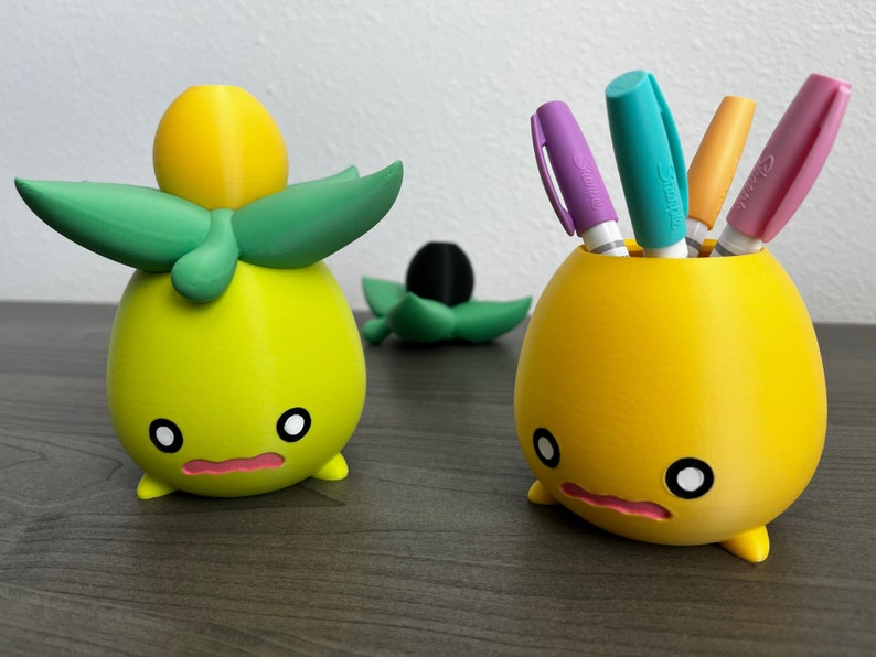May include: Two yellow 3D printed character figurines. One figurine is holding four colorful pens in a cup. The other figurine has green leaves on top of its head.