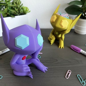 May include: Two stylized figurines, one purple and one yellow, sit on a gray desk. The purple figure has light blue hexagonal eyes and a red gem. The yellow figure holds paperclips. Markers and paperclips are scattered around.