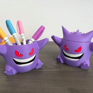 Puede incluir: Dos organizadores de escritorio morados con forma de Gengar. Uno contiene marcadores Sharpie de colores. Los organizadores tienen ojos rojos y amplias sonrisas. Los accesorios de escritorio están diseñados para parecerse al popular personaje de Pokémon.