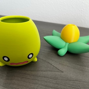 May include: A yellow and green 3D printed planter with a cute face and a flower pot shaped like a yellow bud.