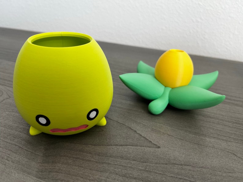 May include: A yellow and green 3D printed planter with a cute face and a flower pot shaped like a yellow bud.