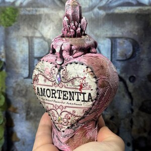 Puede incluir: Una botella de poci&oacute;n Amortentia rosa en forma de coraz&oacute;n con un tap&oacute;n de cristal. La etiqueta dice "Amortentia, The Most Powerful Substance". La botella tiene un aspecto texturizado y envejecido, con adornos decorativos.