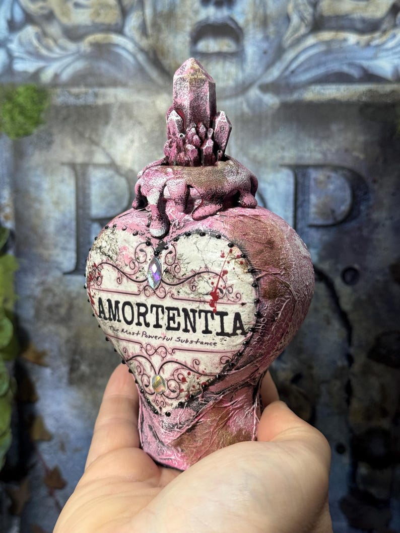 Puede incluir: Una botella de poci&oacute;n Amortentia rosa en forma de coraz&oacute;n con un tap&oacute;n de cristal. La etiqueta dice "Amortentia, The Most Powerful Substance". La botella tiene un aspecto texturizado y envejecido, con adornos decorativos.