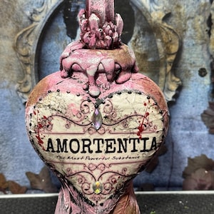 Puede incluir: Una botella de poci&oacute;n Amortentia en forma de coraz&oacute;n con un acabado rosa y dorado. La botella presenta un tap&oacute;n de cristal y el texto "AMORTENTIA: The Most Powerful Substance". Los adornos decorativos incluyen joyas y espirales.