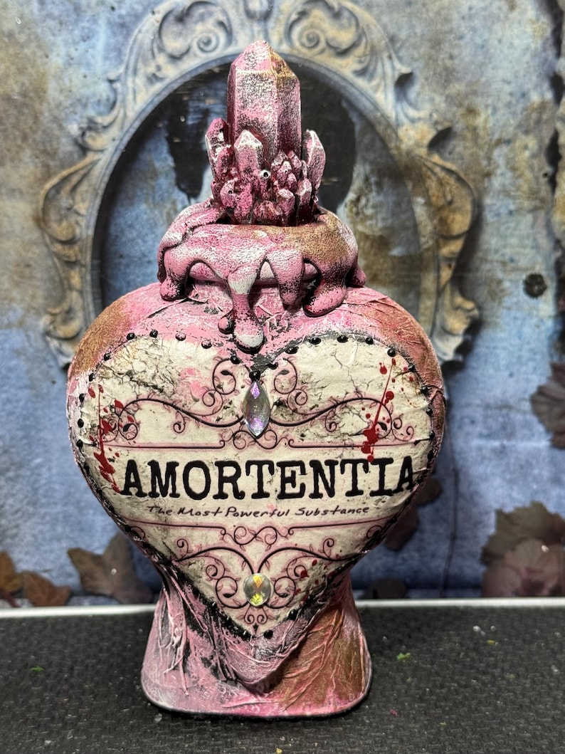 Puede incluir: Una botella de poci&oacute;n Amortentia en forma de coraz&oacute;n con un acabado rosa y dorado. La botella presenta un tap&oacute;n de cristal y el texto "AMORTENTIA: The Most Powerful Substance". Los adornos decorativos incluyen joyas y espirales.