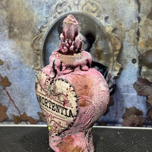 Puede incluir: Una botella rosa en forma de coraz&oacute;n con la palabra "Mortentia" impresa en la parte delantera. La botella tiene un tap&oacute;n en forma de cristal y est&aacute; decorada con detalles dorados. La botella es probablemente un art&iacute;culo decorativo o un accesorio.