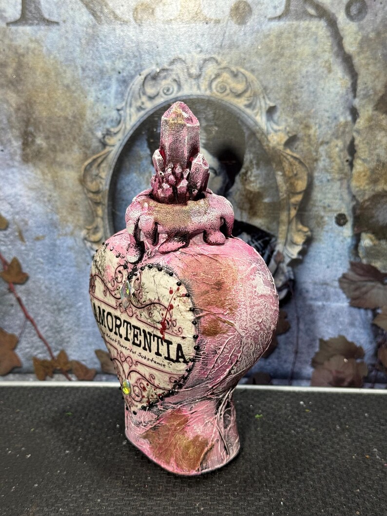 Puede incluir: Una botella rosa en forma de coraz&oacute;n con la palabra "Mortentia" impresa en la parte delantera. La botella tiene un tap&oacute;n en forma de cristal y est&aacute; decorada con detalles dorados. La botella es probablemente un art&iacute;culo decorativo o un accesorio.