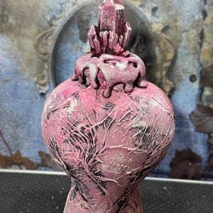 Puede incluir: Un jarr&oacute;n decorativo rosa con una superficie texturizada y una estructura cristalina en la parte superior. El jarr&oacute;n tiene un cuerpo bulboso y una base estrecha. El cristal es de un tono rosa m&aacute;s oscuro y el jarr&oacute;n tiene detalles en blanco y negro.