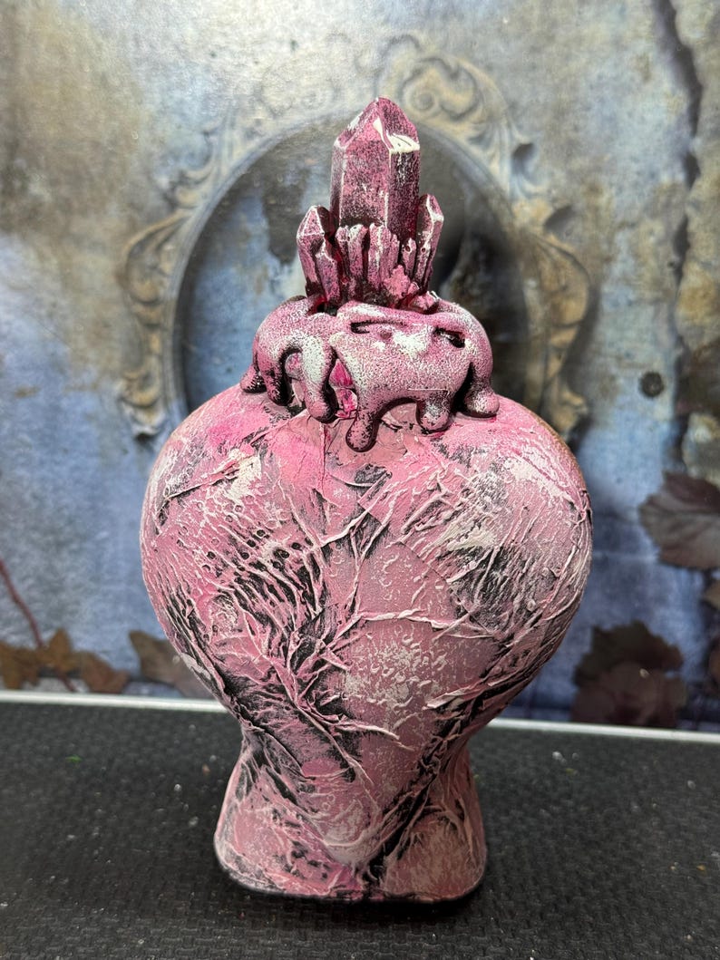 Puede incluir: Un jarr&oacute;n decorativo rosa con una superficie texturizada y una estructura cristalina en la parte superior. El jarr&oacute;n tiene un cuerpo bulboso y una base estrecha. El cristal es de un tono rosa m&aacute;s oscuro y el jarr&oacute;n tiene detalles en blanco y negro.