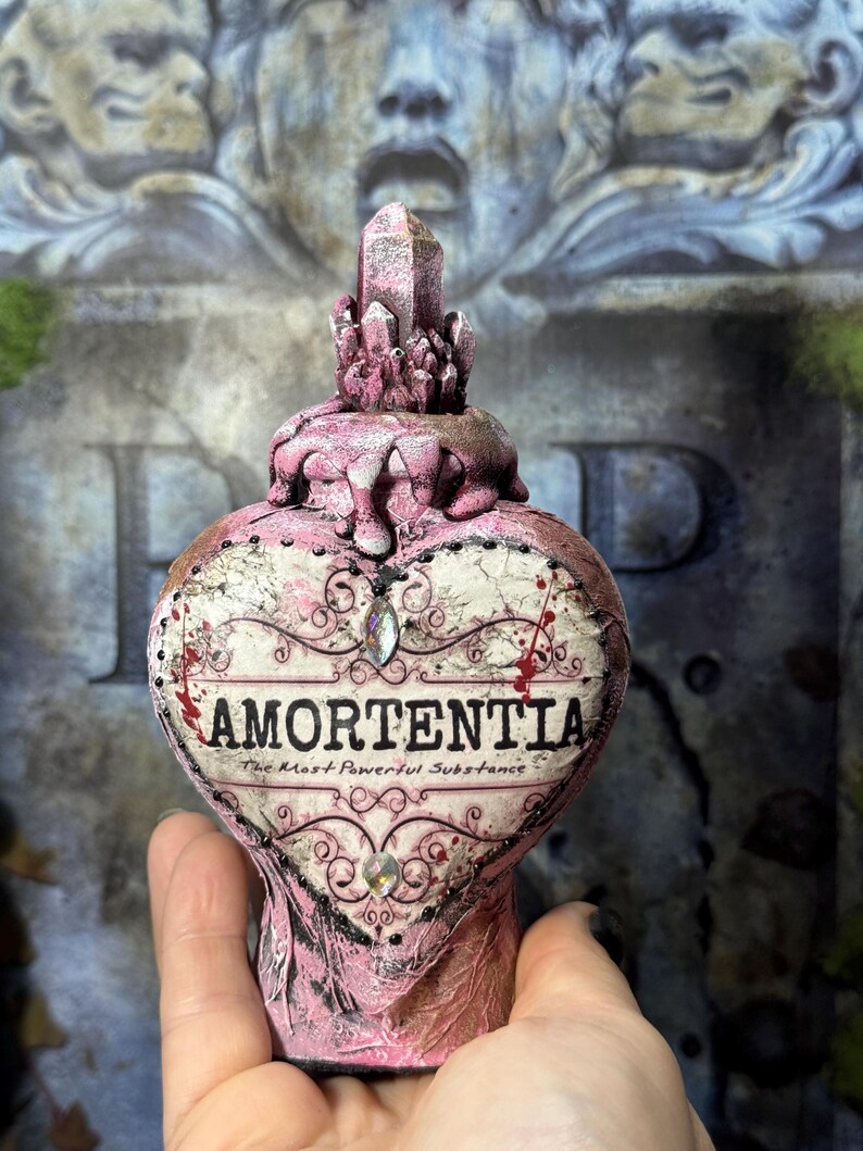 Puede incluir: Una botella rosa en forma de coraz&oacute;n con la palabra "AMORTENTIA" en texto negro. La botella tiene un tap&oacute;n en forma de cristal y est&aacute; decorada con detalles negros y joyas. Tambi&eacute;n es visible el texto "The Most Powerful Substance".