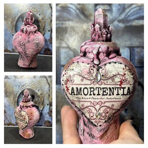 Puede incluir: Una botella de poci&oacute;n Amortentia rosa en forma de coraz&oacute;n con un tap&oacute;n de cristal. La botella tiene una superficie texturizada y las palabras "Amortentia, The Most Powerful Substance" est&aacute;n impresas en la parte delantera. Tambi&eacute;n hay cristales y adornos decorativos.