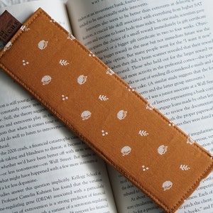 May include: A brown fabric bookmark with a white pattern of small leaves and hedgehogs. The bookmark has a brown leather tab with the word "DECIES" printed on it.