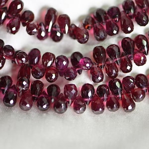 Può includere: Una collana di perle di pietre preziose rosso scuro, ogni perla è sfaccettata e a forma di lacrima. Le perle sono infilate su un filo trasparente e sono disposte in un motivo sciolto e fluido.
