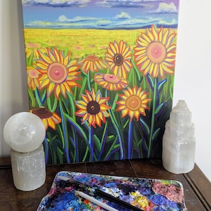 May include: A vibrant painting of sunflowers in a field under a blue sky with fluffy clouds. The artwork features bright yellow, orange, and green hues. A palette with paint and brushes, and selenite crystal sculptures are in the foreground.