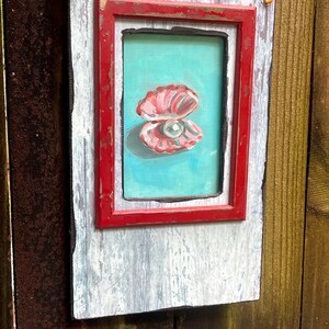 May include: A framed painting of a pink oyster with a pearl on a turquoise background. The painting is set within a red frame, mounted on a distressed white and grey wooden panel. A small loop of twine is attached for hanging.