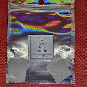 Peut inclure: Un emballage holographique argenté contenant deux zappeurs statiques carrés blancs. L'emballage porte une étiquette indiquant "J&L's Scented Static Zappers made with love."