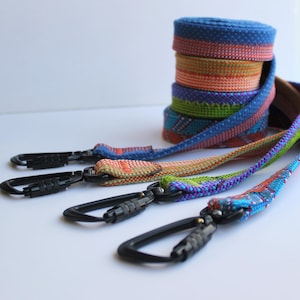 May include: A stack of colorful woven fabric straps featuring various geometric patterns in shades of blue, orange, green, and purple. Each strap is attached to a black metal carabiner clip with a screw-lock mechanism for secure fastening.