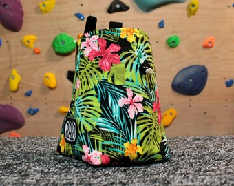 May include: A climbing chalk bag with a tropical floral pattern of pink, yellow, and green flowers and leaves on a black background. The bag has a black drawstring closure and a small logo patch.