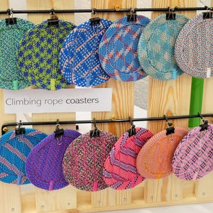 May include: A collection of twelve round coasters made from woven climbing rope, displayed on a wooden rack with black metal clips. The coasters feature various patterns and colors, including blue, green, pink, and orange. A sign reads Climbing rope coasters.