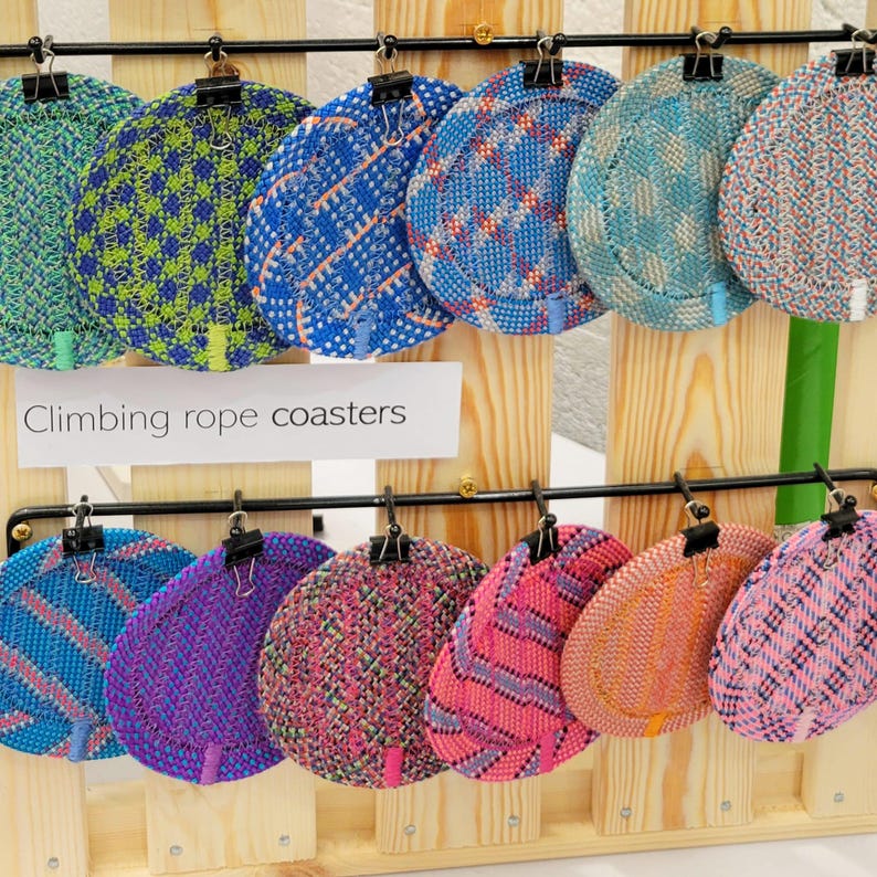 May include: A collection of twelve round coasters made from woven climbing rope, displayed on a wooden rack with black metal clips. The coasters feature various patterns and colors, including blue, green, pink, and orange. A sign reads Climbing rope coasters.
