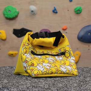 Pode incluir: Uma bolsa de giz de escalada amarela com fecho de correr e uma correia preta. A bolsa &eacute; coberta com um padr&atilde;o de faces de patos amarelos.