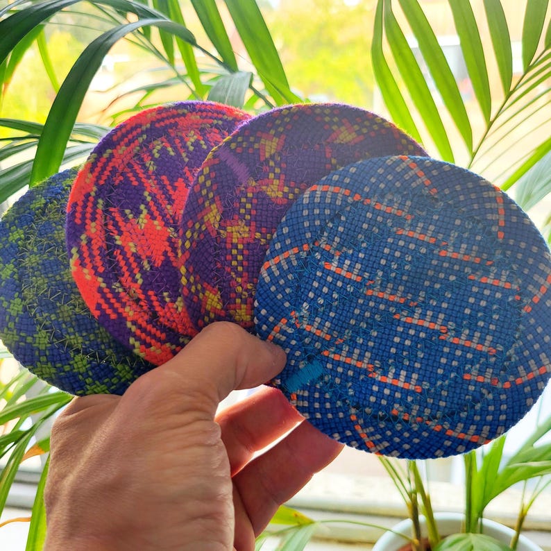 May include: Four circular coasters featuring woven fabric patterns in distinct color combinations. The set includes a green and blue, a purple and orange, a purple and yellow, and a blue and orange design, all with visible stitched edges.