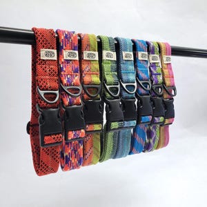 May include: A row of eight woven dog collars in various colors including red, orange, green, blue, and purple. Each collar features a black plastic side-release buckle, a black metal D-ring, and a small white fabric tag with the text RAKD.