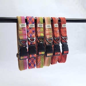 May include: Six woven fabric pet collars hanging on a black rod. Each collar features a unique multicolored pattern in shades of pink, orange, green, and red, with a black plastic side-release buckle, a metal D-ring, and a small white brand tag.
