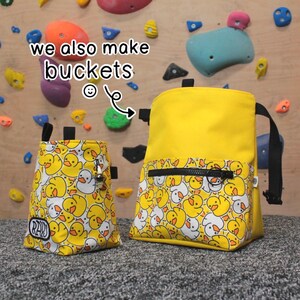May include: Two yellow climbing chalk bags with a duck pattern. The larger bag has a zippered pocket. The smaller bag has the word "RAD" printed on it. The text "we also make buckets" is written on the image.