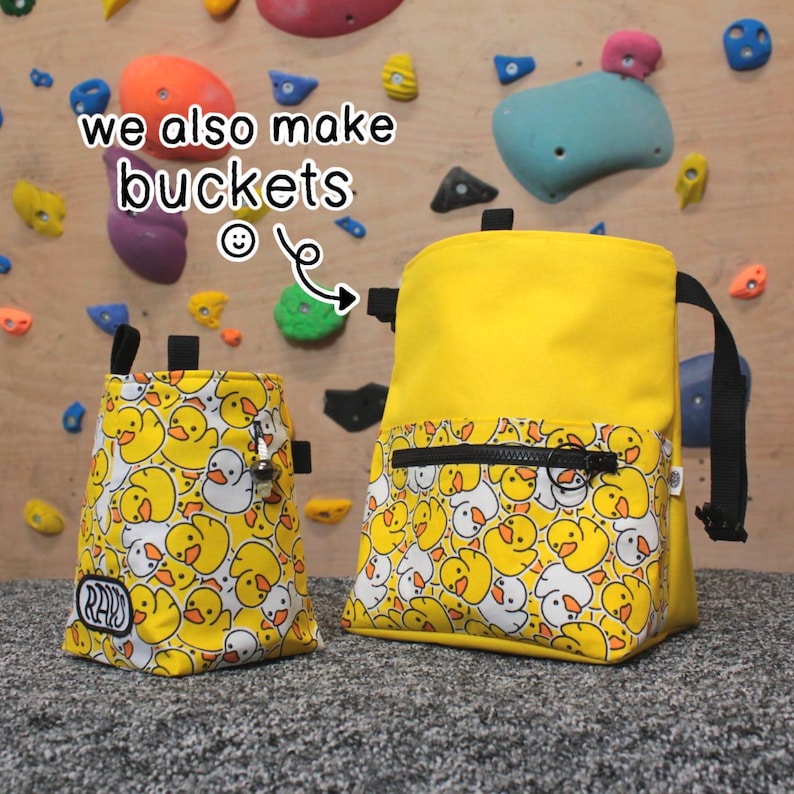 May include: Two yellow climbing chalk bags with a duck pattern. The larger bag has a zippered pocket. The smaller bag has the word "RAD" printed on it. The text "we also make buckets" is written on the image.