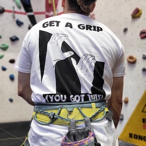 Può includere: Maglietta bianca con la scritta "GET A GRIP" e "(YOU GOT THIS)" stampata in nero. La maglietta presenta una grafica di due mani che escono da un quadrato nero. È visibile un'imbracatura da arrampicata gialla e grigia.