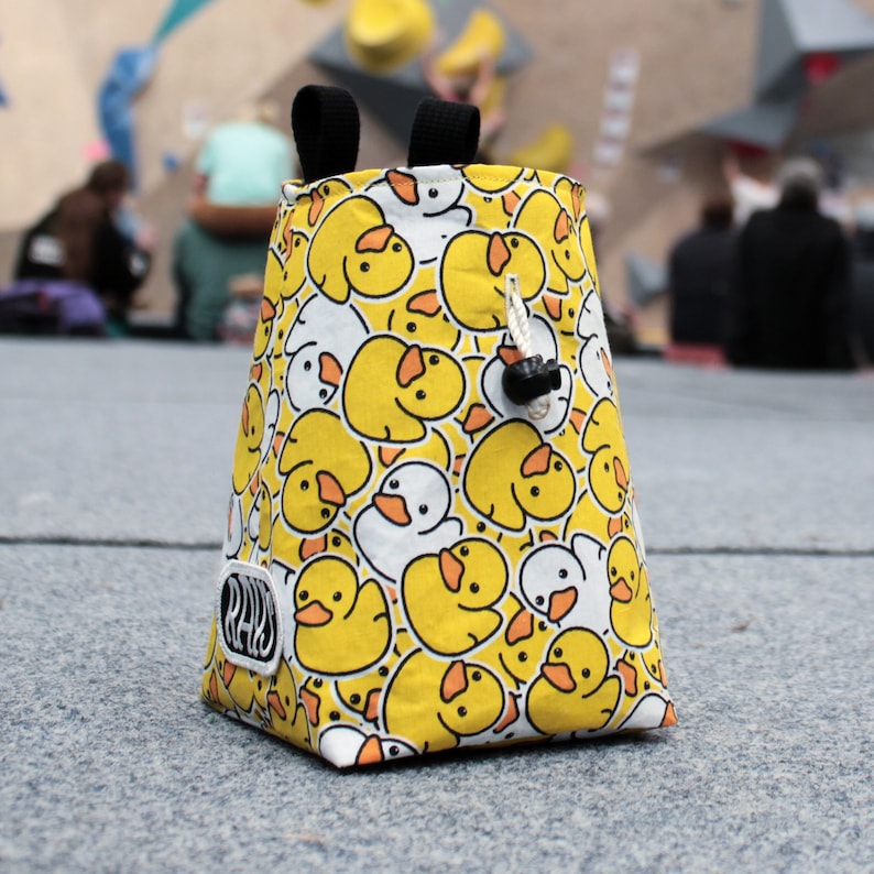May include: A yellow and white climbing chalk bag with a rubber ducky pattern. The bag has a drawstring closure and a black label with the word "RAD" on it.