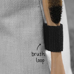 May include: A close-up of a black brush loop attached to a white canvas bag. The loop is designed to hold a brush or other tool securely.