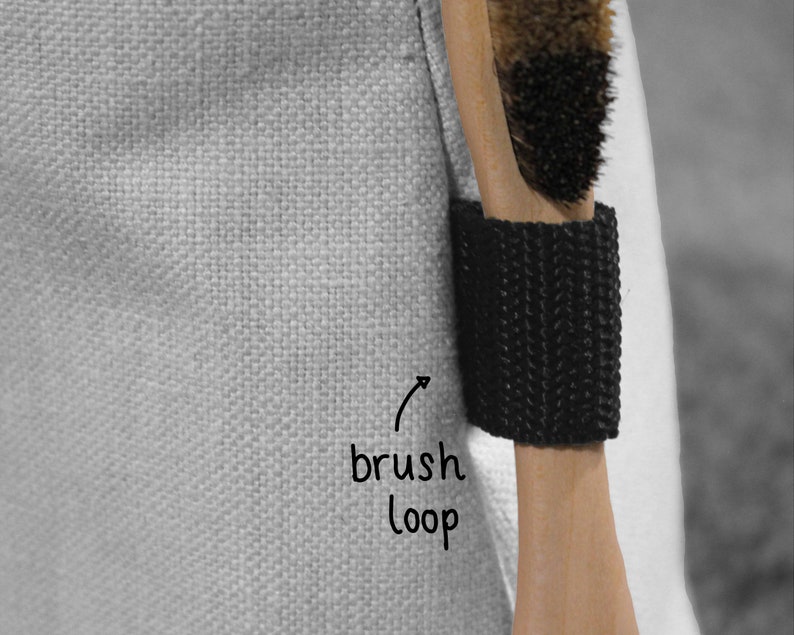 May include: A close-up of a black brush loop attached to a white canvas bag. The loop is designed to hold a brush or other tool securely.
