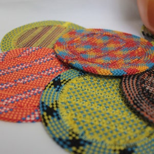 May include: A collection of five circular coasters featuring intricate woven patterns in vibrant colors including orange, yellow, blue, and black. Each coaster displays a unique geometric design created by the interlacing of thin, textured strands.