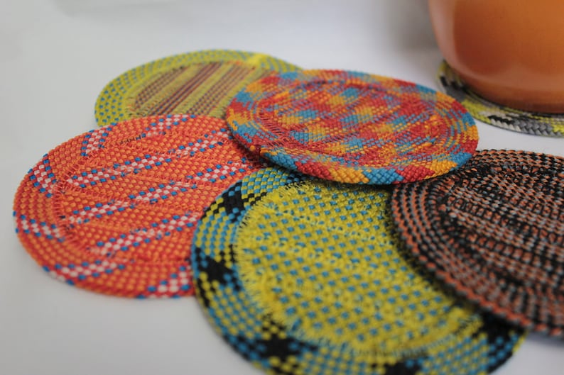 May include: A collection of five circular coasters featuring intricate woven patterns in vibrant colors including orange, yellow, blue, and black. Each coaster displays a unique geometric design created by the interlacing of thin, textured strands.