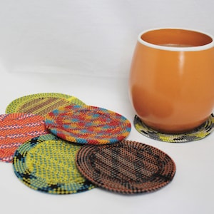 May include: A round orange mug with a white rim sits on a circular coaster with a grey and yellow pattern. Four additional round coasters are arranged nearby, featuring woven designs in vibrant colors including orange, yellow, blue, and black.